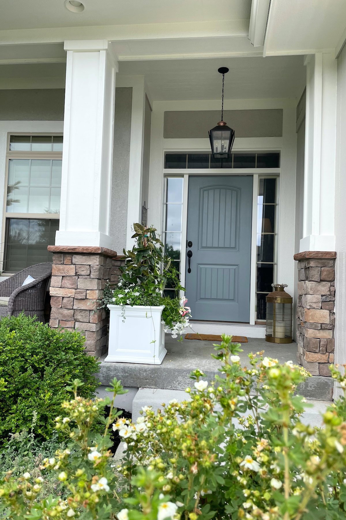 A house entrance with a light blue door, flanked by white columns and stone accents. A white planter with greenery sits near the door, painted in Benjamin Moore Brewster Gray, and a hanging lantern light fixture is above it. Bushes in the foreground.