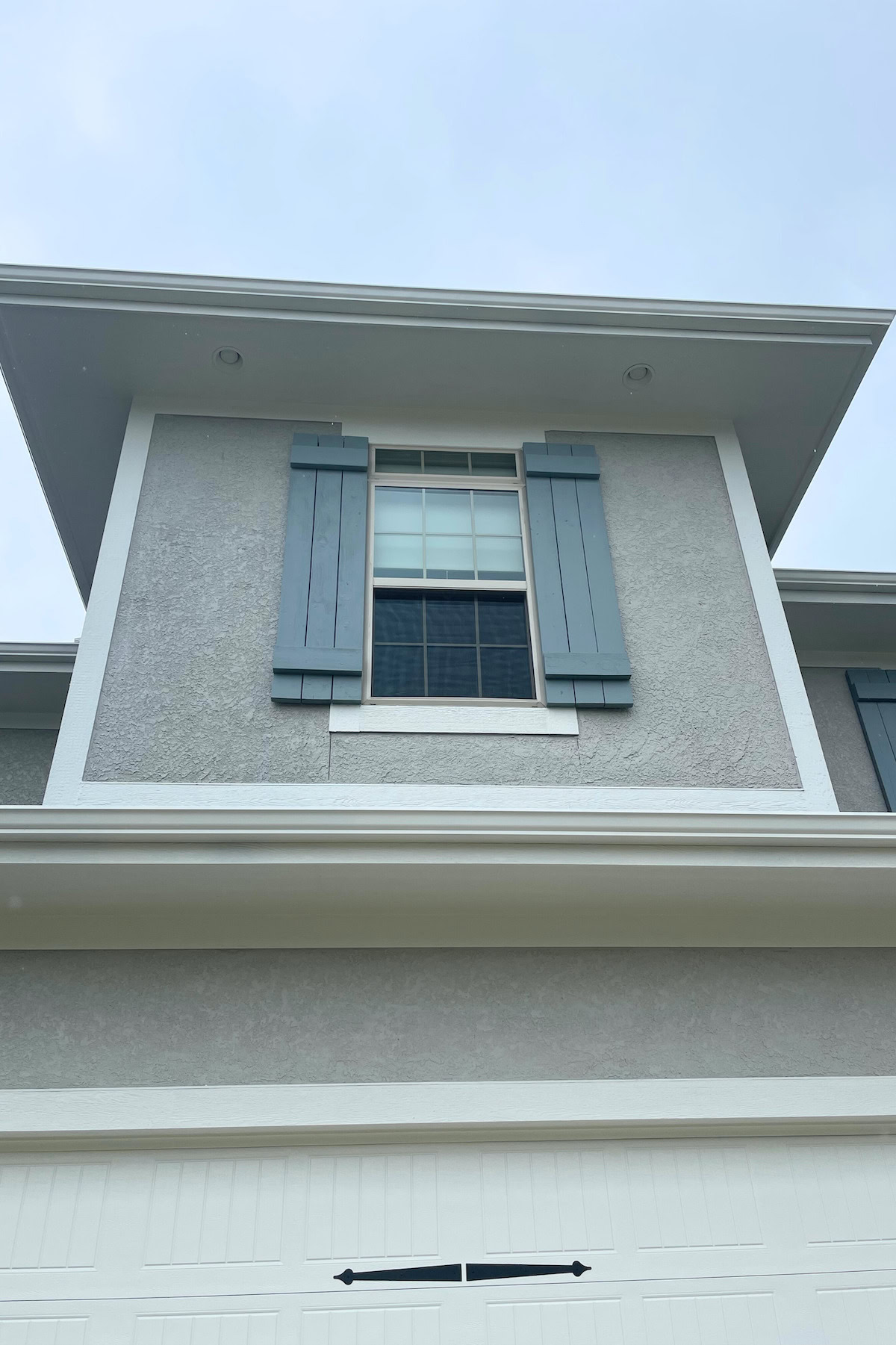 Close-up view of a two-story house's upper window with blue shutters. The Benjamin Moore Brewster Gray exterior and white trim frame the window, and part of a white garage door is visible at the bottom.