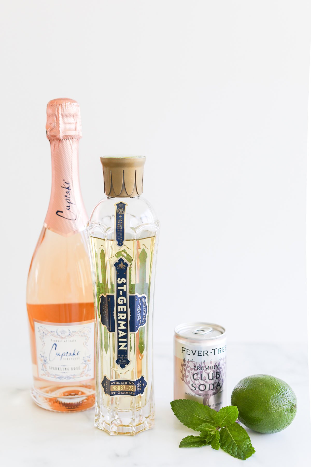 A bottle of rosé sparkling wine, a bottle of St-Germain liqueur, a can of club soda, a lime, and mint leaves arranged on a white surface.