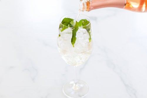 A glass of rosé spritz is being topped off with a bottle of rosé poured from a bottle with mint leaves on top, placed on a white marble surface.