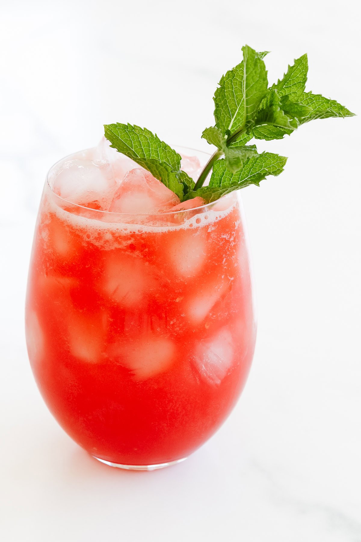 A cold, red watermelon juice with ice cubes, garnished with a sprig of fresh mint in a clear glass on a white surface.