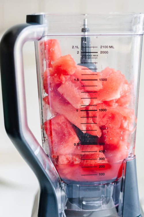 A blender container filled with cut watermelon pieces and some water, with measurement markings visible on the side.