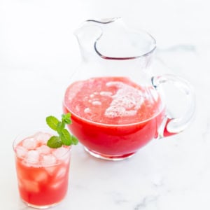 A glass pitcher filled with watermelon juice and a glass of the same juice with ice cubes and a mint garnish, both placed on a white surface.