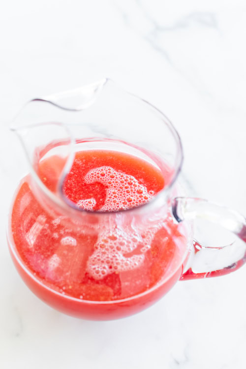A clear glass pitcher filled with frothy watermelon juice, placed on a white surface.