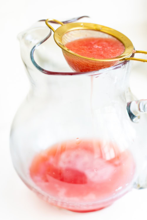 A clear glass pitcher with a small amount of watermelon juice at the bottom, being strained through a fine mesh sieve with red pulp collected in the sieve.