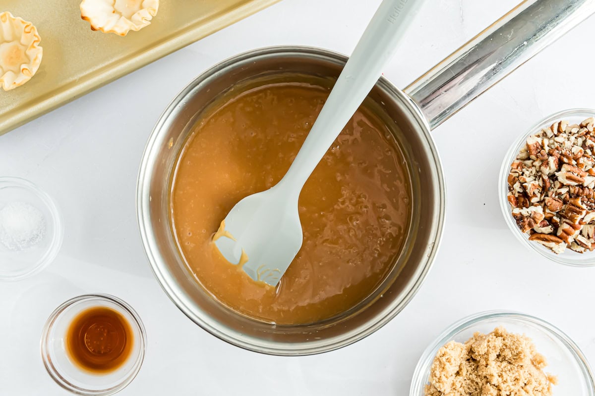 A saucepan filled with caramel sauce is placed on a white surface, surrounded by small bowls containing pecans, brown sugar, vanilla extract, and a pinch of salt.