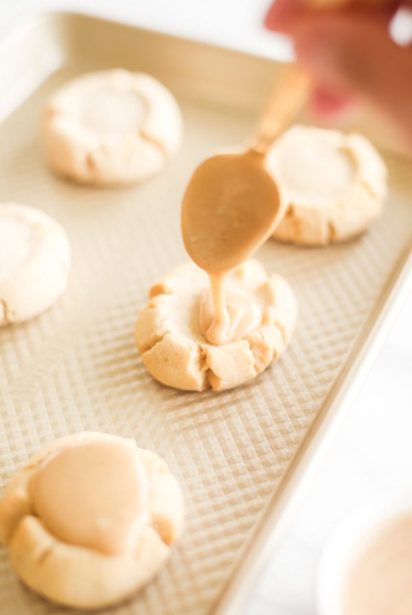 A hand is using a spoon to drizzle caramel onto cookies on a baking sheet.