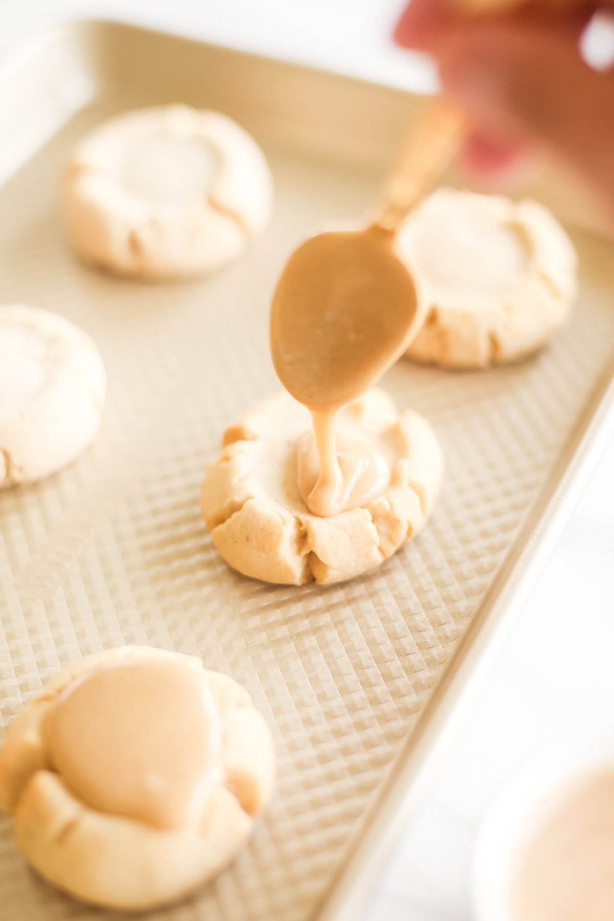 A hand is using a spoon to drizzle caramel onto cookies on a baking sheet.