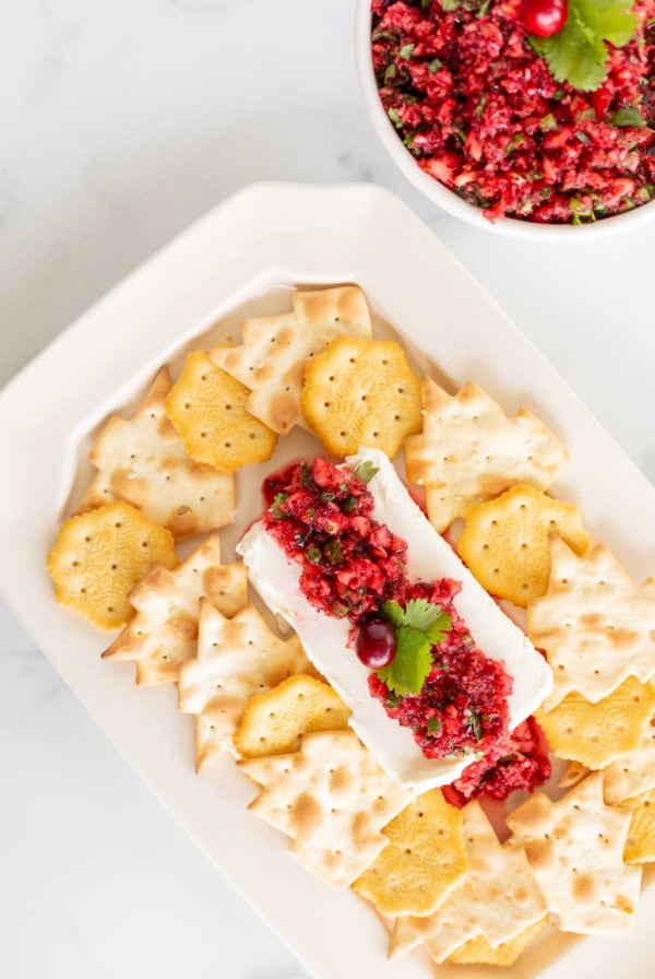 A plate of crackers arranged around a block of cream cheese topped with a red, finely chopped mixture of cranberry salsa