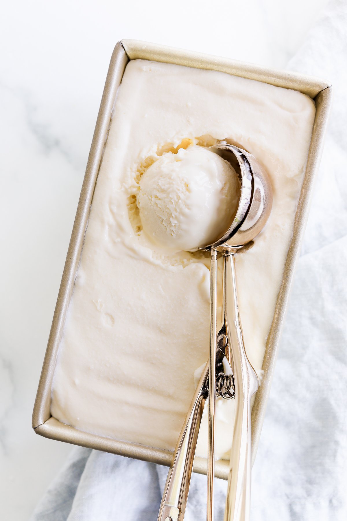 A rectangular container of Gooey Butter Cake Ice Cream with a metal scooper holding a creamy scoop.