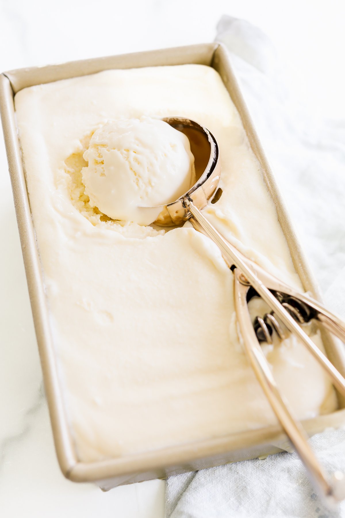 A metal ice cream scoop rests in a rectangular container of Gooey Butter Cake ice cream, with one scoop of the rich treat inside the scooper. A white cloth is partially visible beside the container.