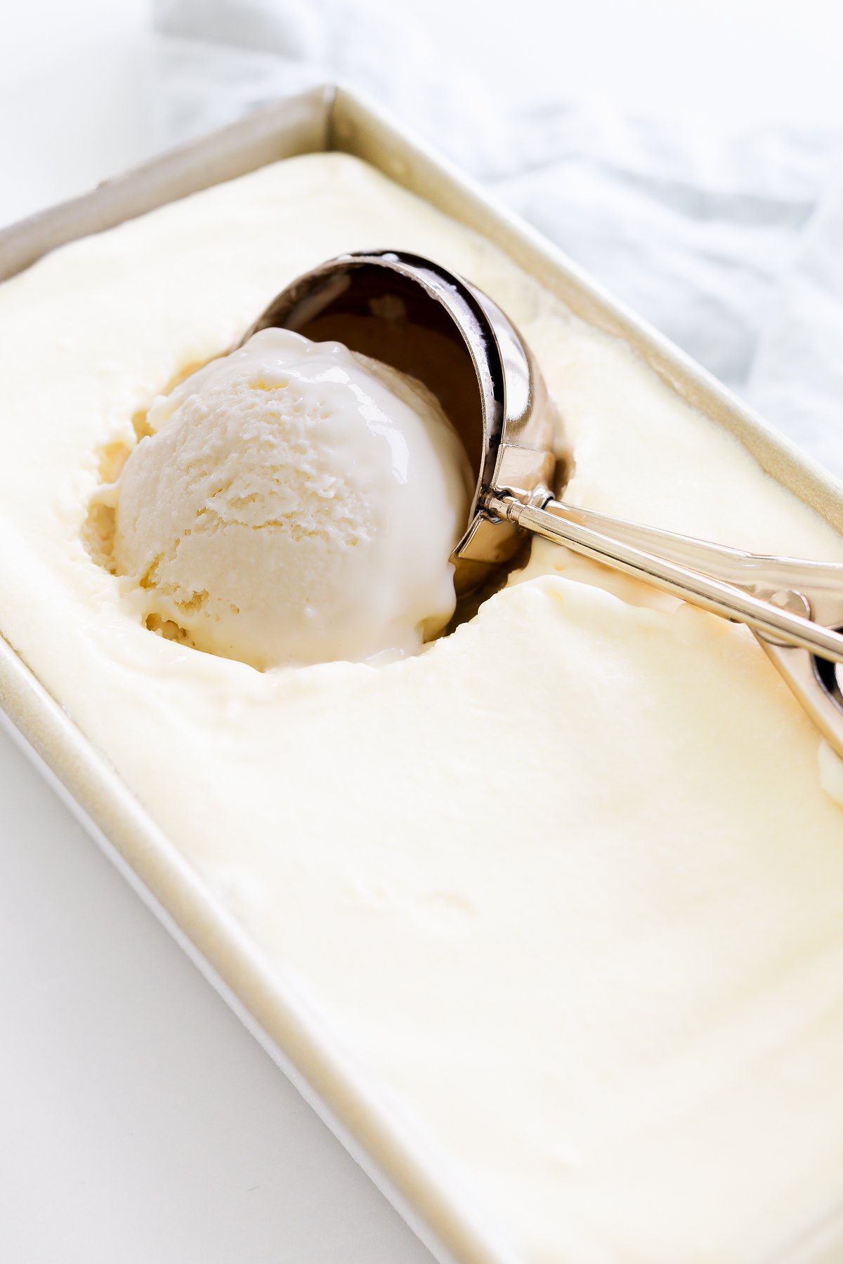 An ice cream scoop is in the process of scooping Gooey Butter Cake Ice Cream from a rectangular metal container. The creamy texture of the ice cream is visible.