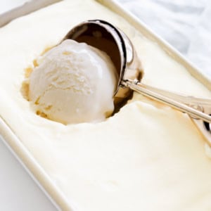 A metal ice cream scooper is scooping a ball of Gooey Butter Cake Ice Cream from a rectangular container.