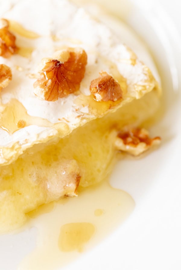 Close-up of a baked brie cheese topped with honey and walnuts, melting on a white plate.