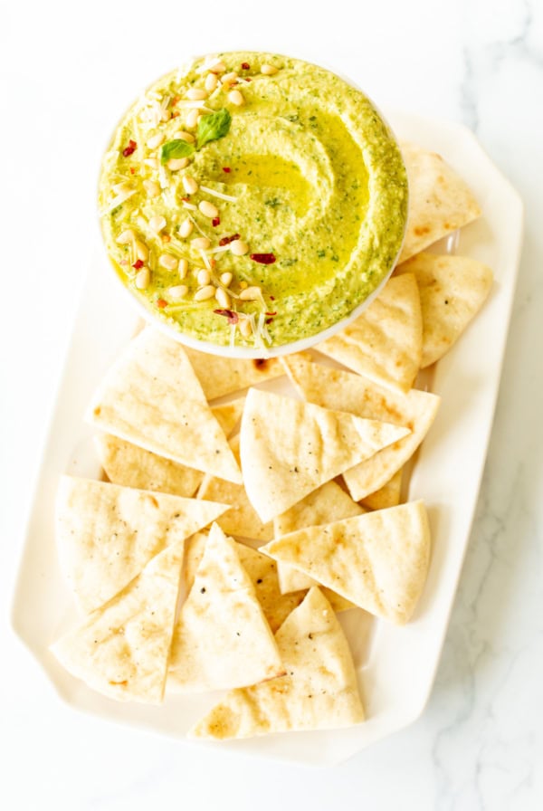 A bowl of green hummus blended with pesto and garnished with pine nuts and microgreens is served on a rectangular white plate with triangular pieces of pita bread.