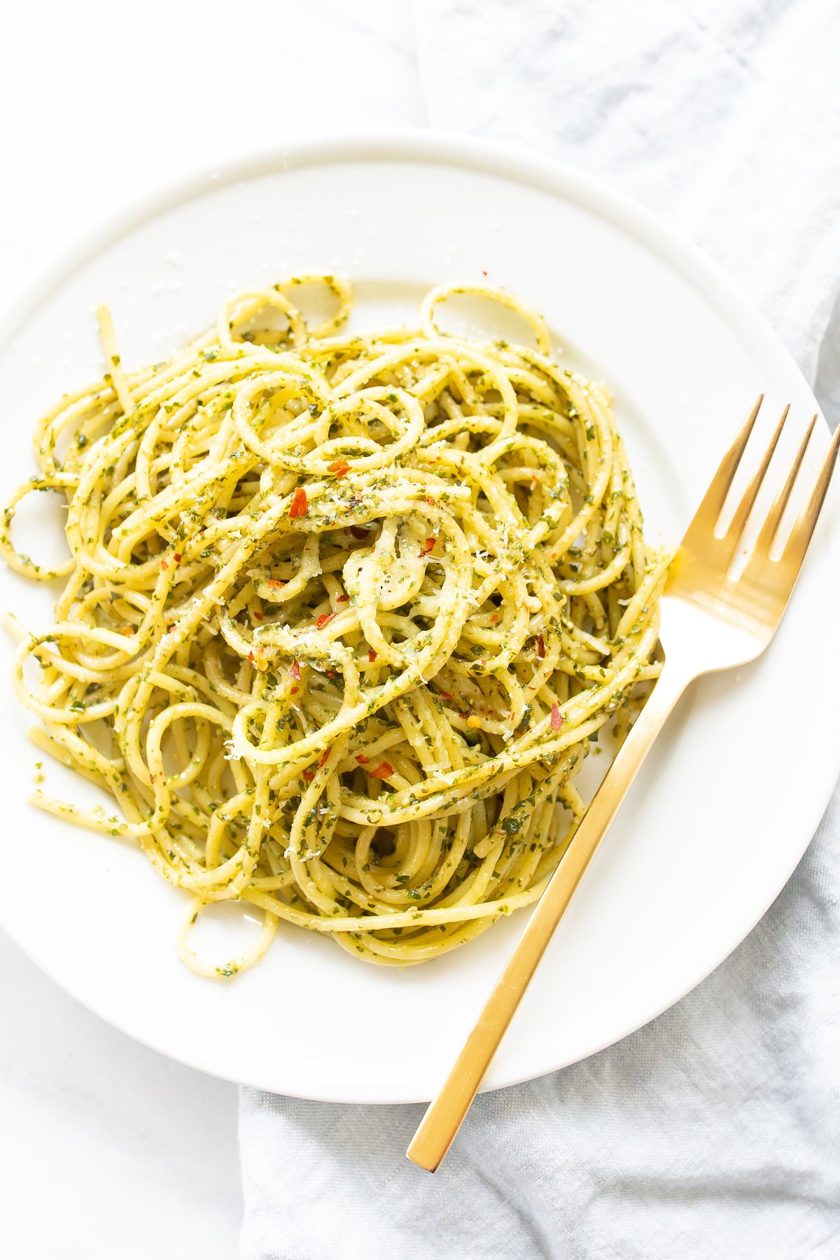 A white plate holds a serving of spaghetti tossed in green pesto sauce, garnished with red pepper flakes. A gold fork rests elegantly to the right of the plate. Perfect for those exploring recipes with pesto, this dish offers a vibrant and flavorful experience.