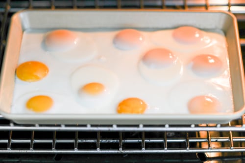 A sheet pan filled with eight sunny-side-up eggs is placed in an oven.