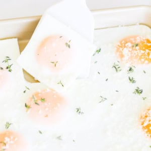 A white spatula lifts a square of sheet pan eggs topped with grated cheese and fresh herbs from a baking tray.