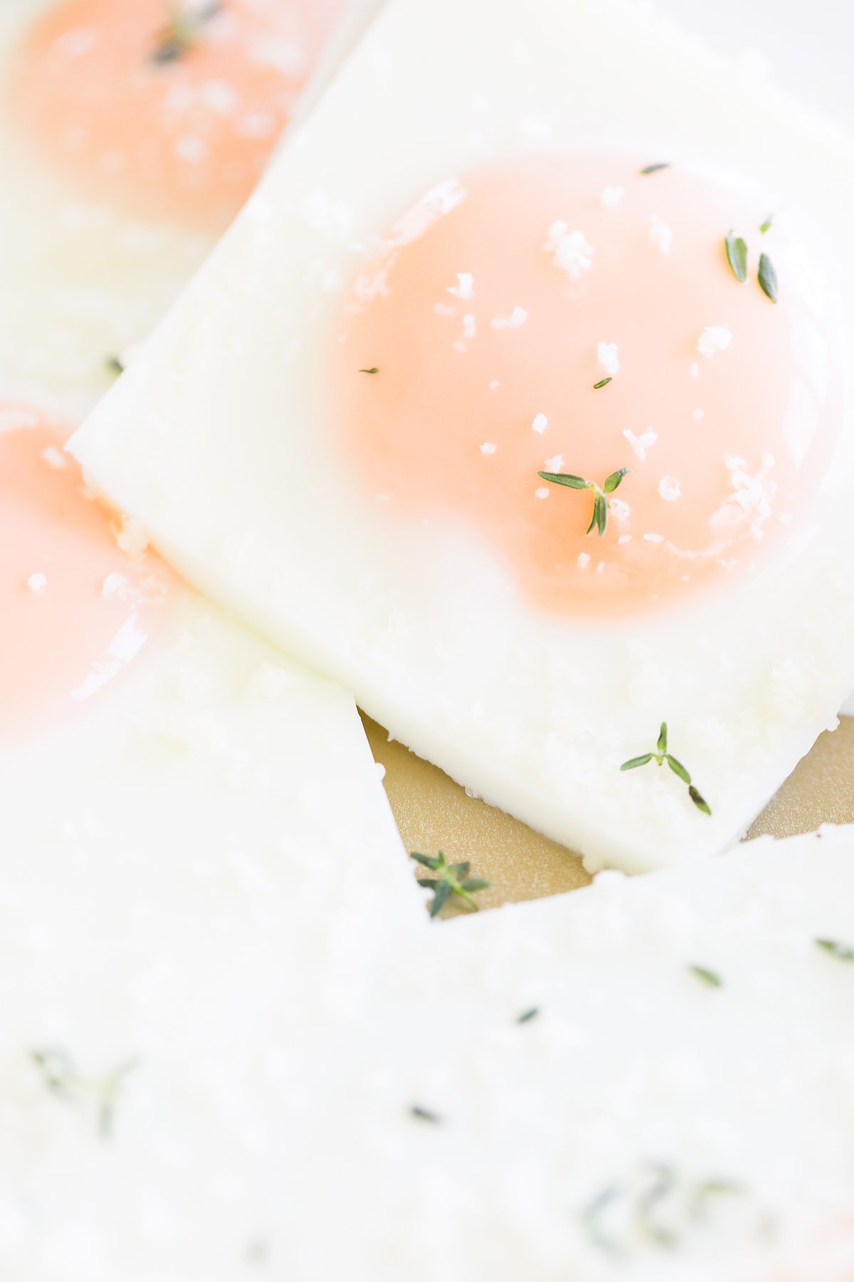 Close-up of sheet pan eggs sunny-side-up, garnished with salt flakes and fresh thyme sprigs, neatly arranged on a white surface.