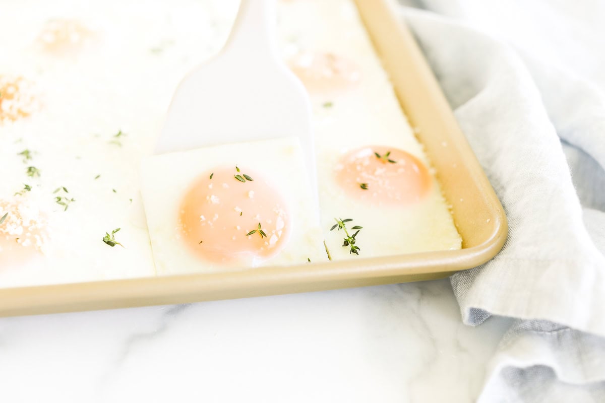 A baking tray with Sheet Pan Eggs garnished with herbs. A white spatula is lifting one egg. The tray is placed on a white marble surface with a light grey cloth nearby.