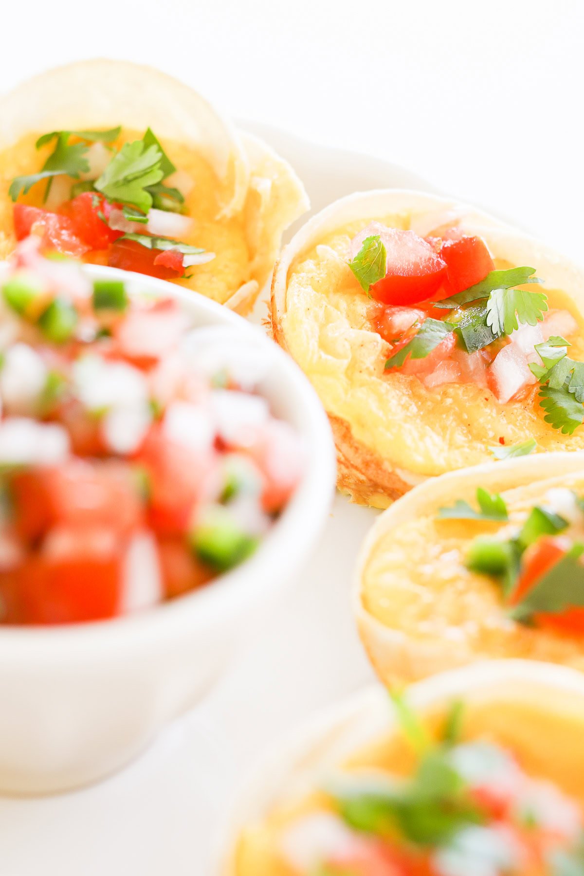 Close-up of Mexican Breakfast Cups garnished with herbs and diced tomatoes, accompanied by a bowl of fresh salsa on a white plate.