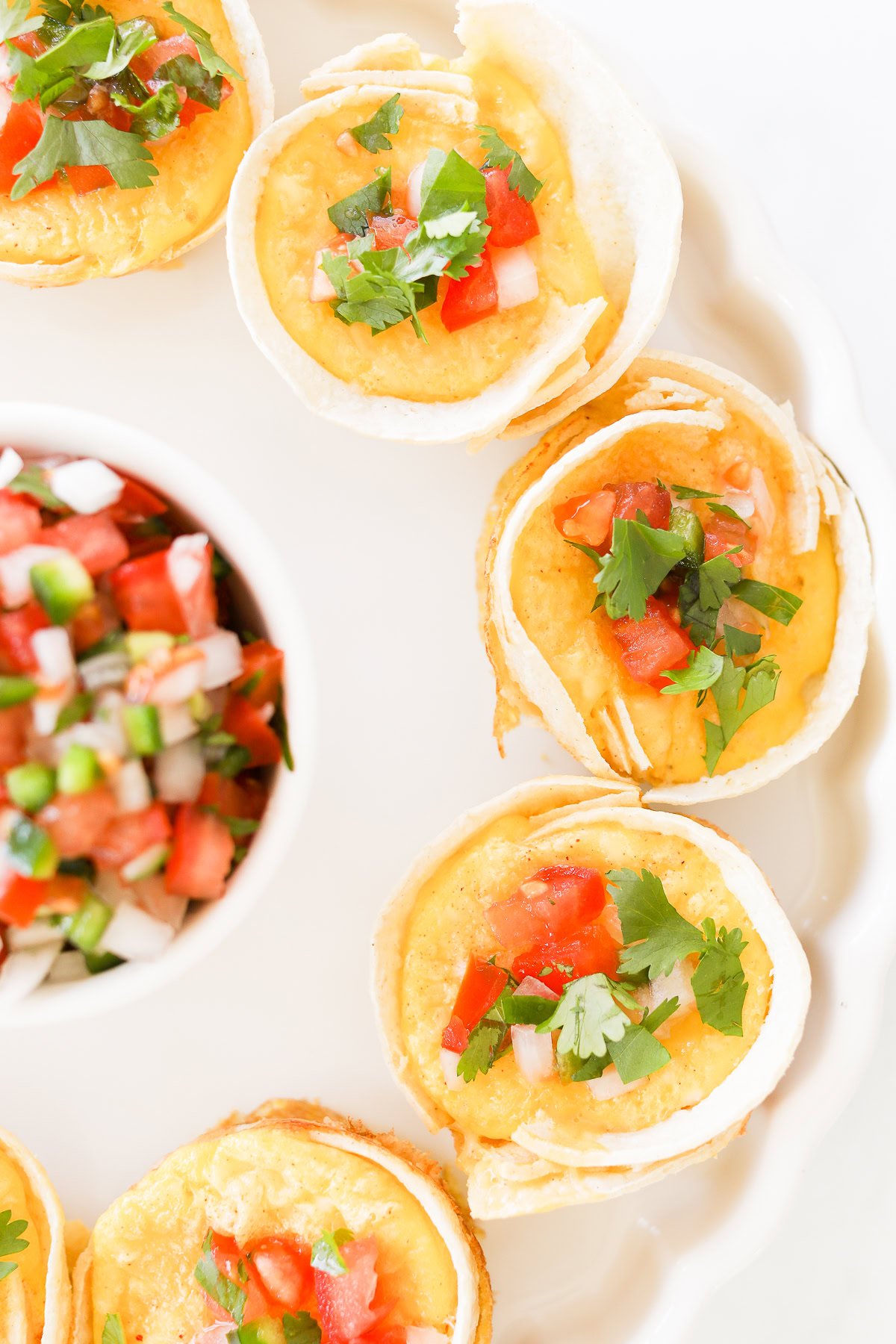 A plate of mini Mexican Breakfast Cups filled with melted cheese, diced tomatoes, onions, and cilantro, arranged in a circle with a small bowl of salsa in the center.