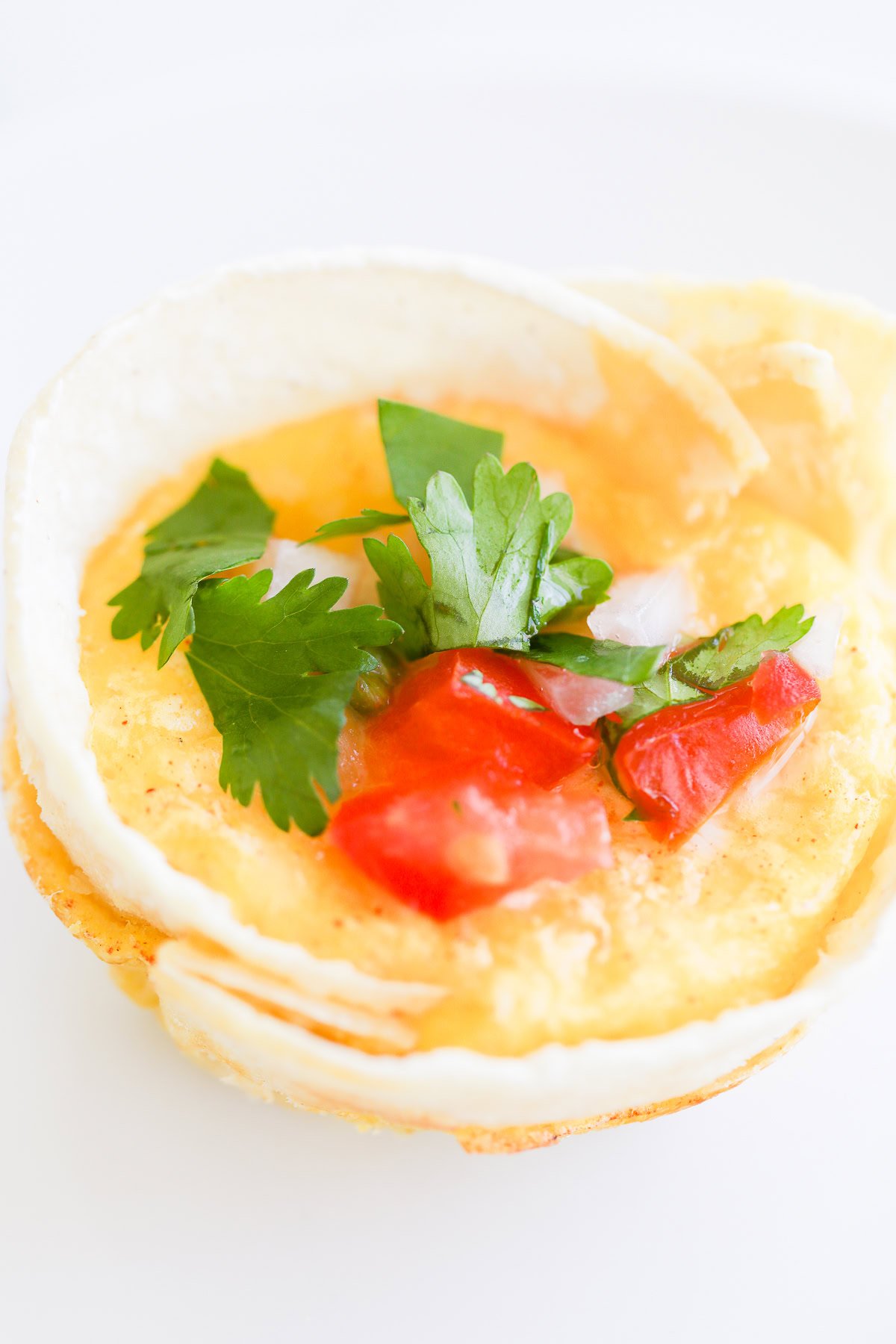 A close-up of a small savory dish: Mexican Breakfast Cups featuring scrambled eggs, topped with parsley, diced tomatoes, and onions, all served in a white edible shell.