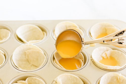 A scoop of liquid filling is being poured into tortilla cups in a muffin tin, creating delightful Mexican Breakfast Cups.