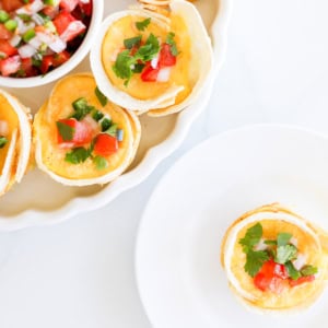 Mexican Breakfast Cups: Small tortilla egg cups filled with eggs, topped with chopped tomatoes, jalapenos, and cilantro, served on a white plate with a side of salsa.