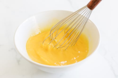 A wire whisk stirring a yellow egg mixture in a white bowl on a white marble surface, perfect for making delicious Mexican Breakfast Cups.