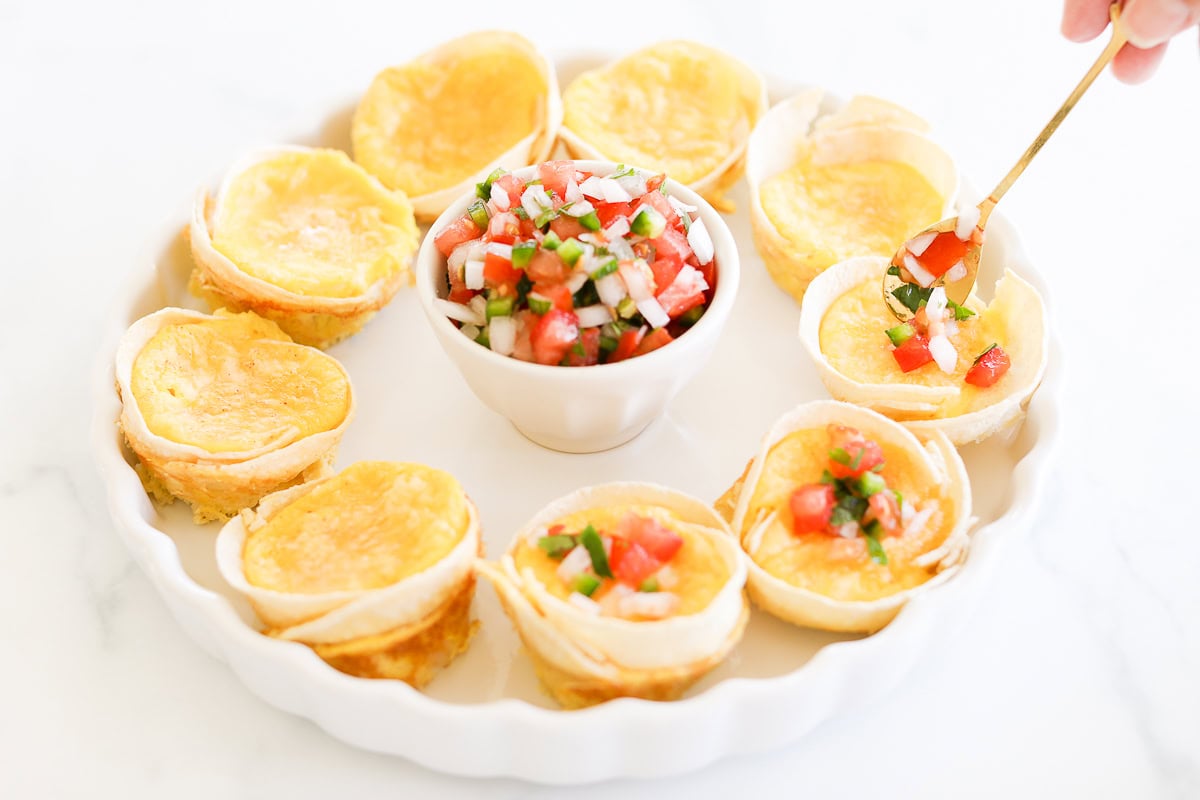 A white plate with small Mexican Breakfast Cups filled with melted cheese arranged in a circle around a bowl of diced tomato, onion, and cilantro salsa. A hand uses a spoon to scoop salsa into one of the tortilla cups.