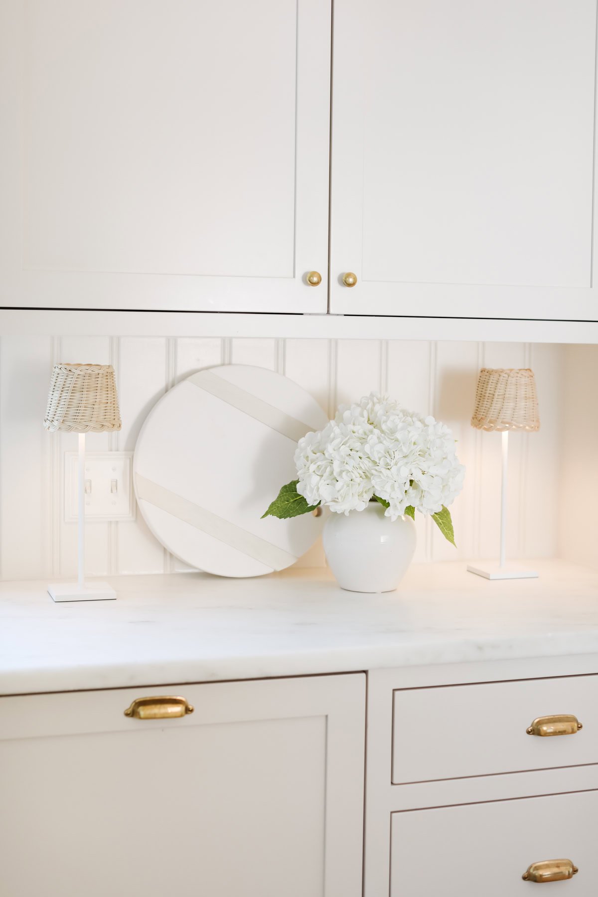 A white kitchen countertop showcases a vase of white flowers, a round decorative tray, and two small lamps with woven shades. Cabinets with brass knobs are above and below, presenting an inviting space that's perfect for styling your home this Prime Day.