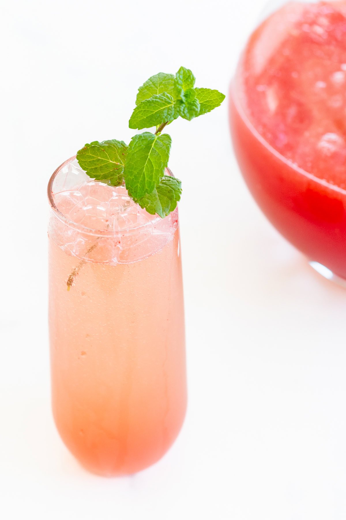 A tall glass filled with a pink-hued watermelon mimosa, garnished with a sprig of fresh mint. A large bowl, partially visible, contains more of the same beverage in the background.