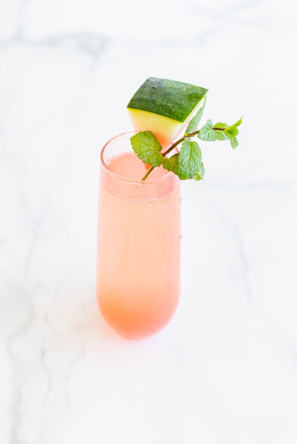 A tall glass of pink watermelon mimosa garnished with a cube of fresh watermelon and a sprig of mint rests on a white marble surface.