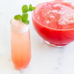 A tall glass filled with a pink-hued watermelon mimosa, garnished with a sprig of fresh mint. A large bowl, partially visible, contains more of the same beverage in the background.