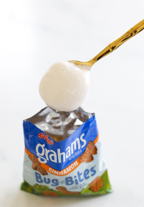 A gold spoon holds a white marshmallow over an open bag of Kellogg's Graham Crackers Bug Bites, cinnamon flavor, creating the perfect setup for walking s'mores.