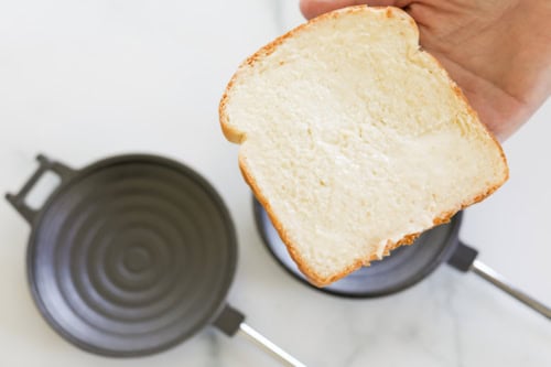 A hand holds a slice of bread spread with butter above a stovetop pan, reminiscent of rustic campfire pizza simplicity.