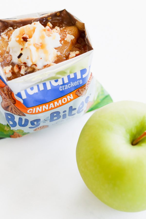 An open apple pie in a bag topped with whipped cream and nuts sits enticingly next to a crisp green apple on a white surface.