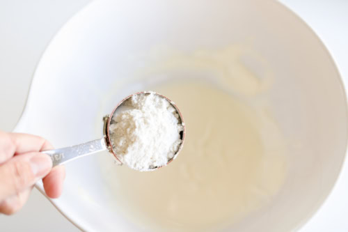 A hand carefully holds a measuring cup filled with flour above a mixing bowl, preparing to create the perfect birthday cake popcorn treat.