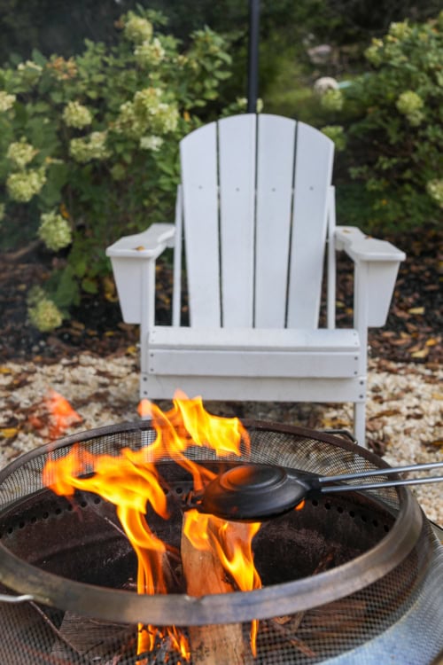 A white Adirondack chair near a fire pit with flames, a sandwich press, and the aroma of Campfire Pizza wafting through the air, all surrounded by lush greenery.