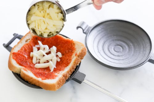 A hand sprinkles shredded cheese onto a slice of bread with tomato sauce, creating a campfire pizza in a round sandwich press on a white surface.