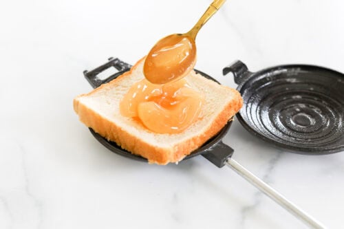 A spoonful of marmalade being spread on a slice of white bread placed in a black sandwich maker on a white surface.