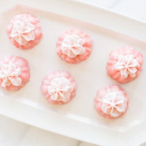 Six pink pastries adorned with elegant champagne frosting are arranged on a rectangular white plate, each topped with delicate white decorations.