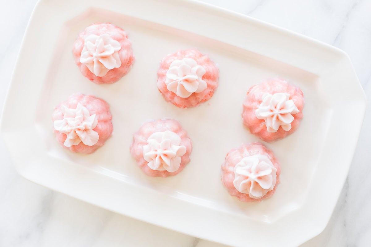 Six pink pastries adorned with elegant champagne frosting are arranged on a rectangular white plate, each topped with delicate white decorations.