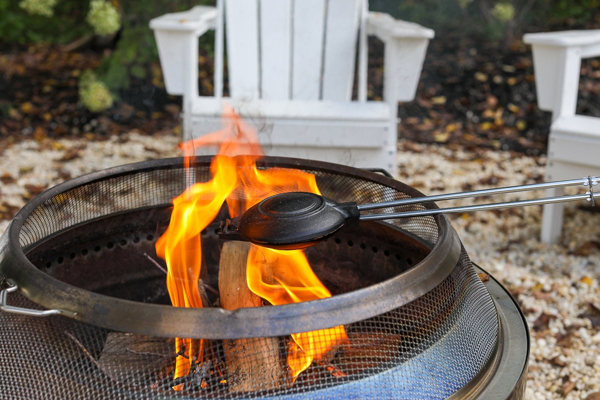 A pie iron sizzles with campfire pizza, flames flickering in the background, while two white Adirondack chairs invite relaxation on the gravel ground.