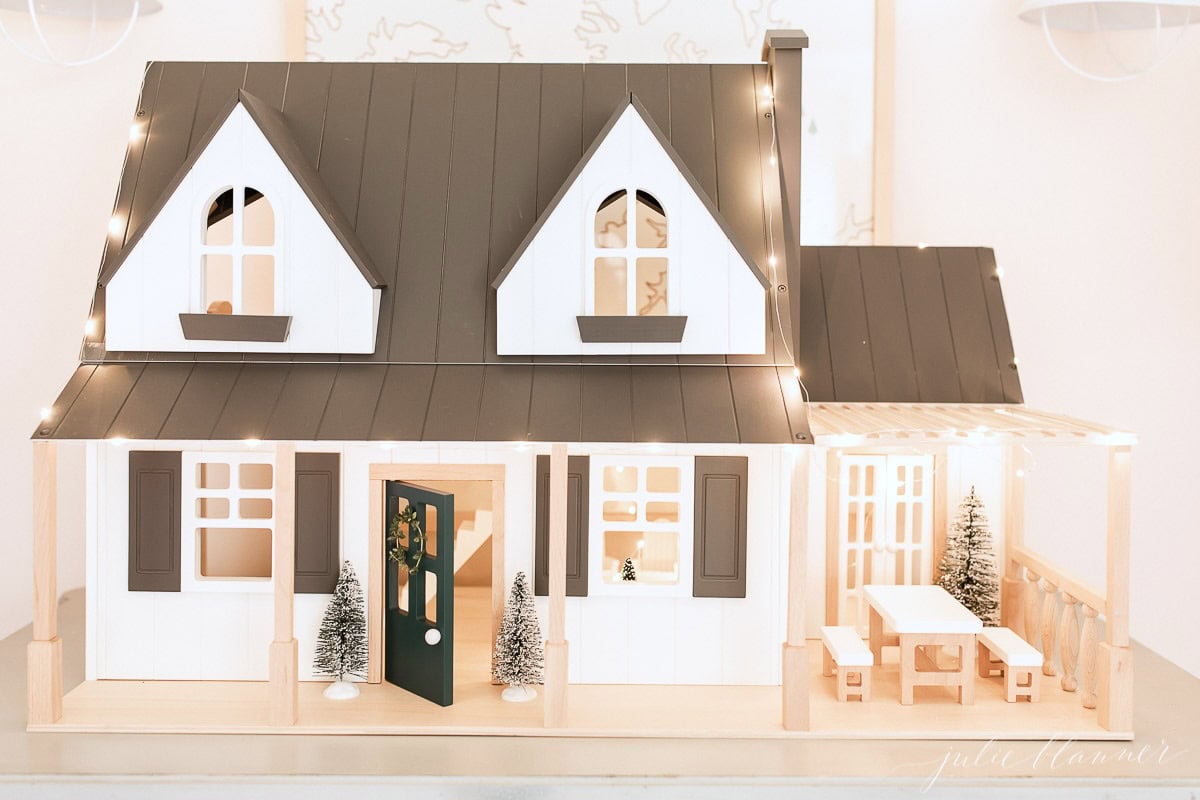 A miniature model of a white and brown house with lit windows, two small trees at the front, and a porch with a table and bench, perfect for those looking to capture the coziness of Prime Day.