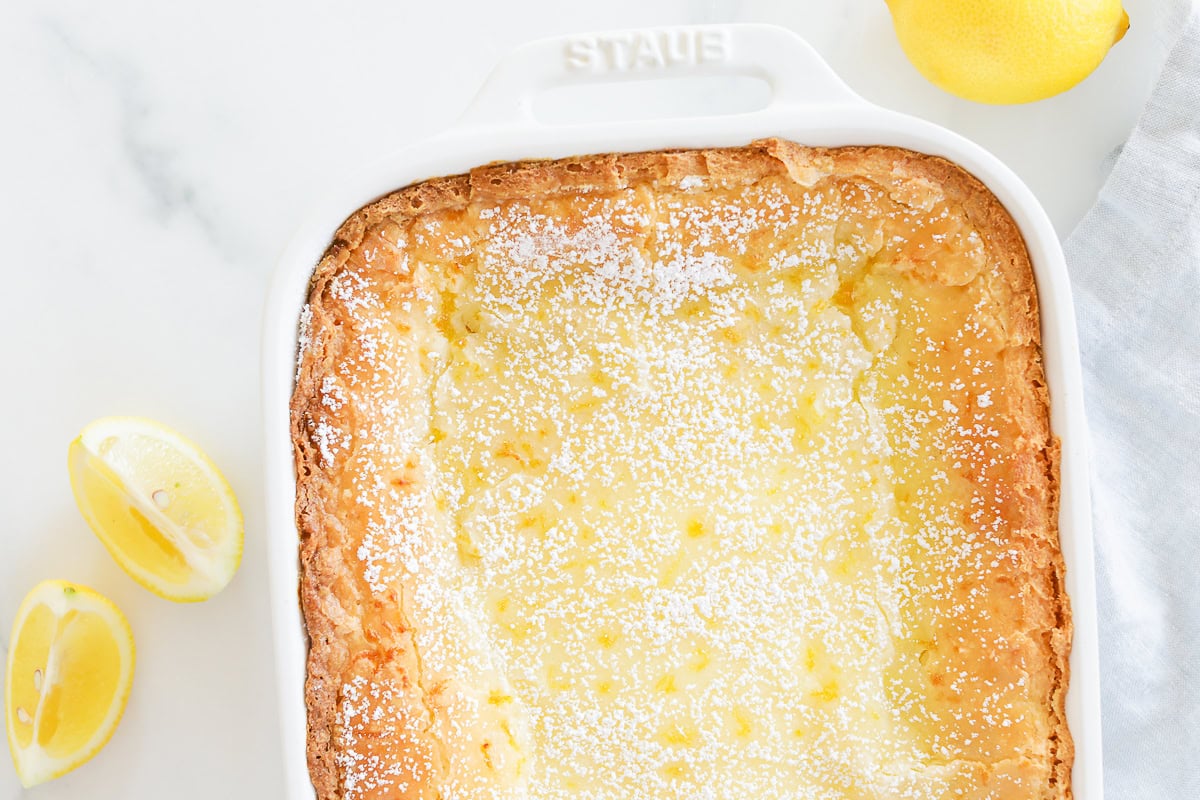 A rectangular Lemon Gooey Butter Cake, dusted with powdered sugar, sits elegantly in a white baking dish. Two lemon halves rest on the marble surface beside it.