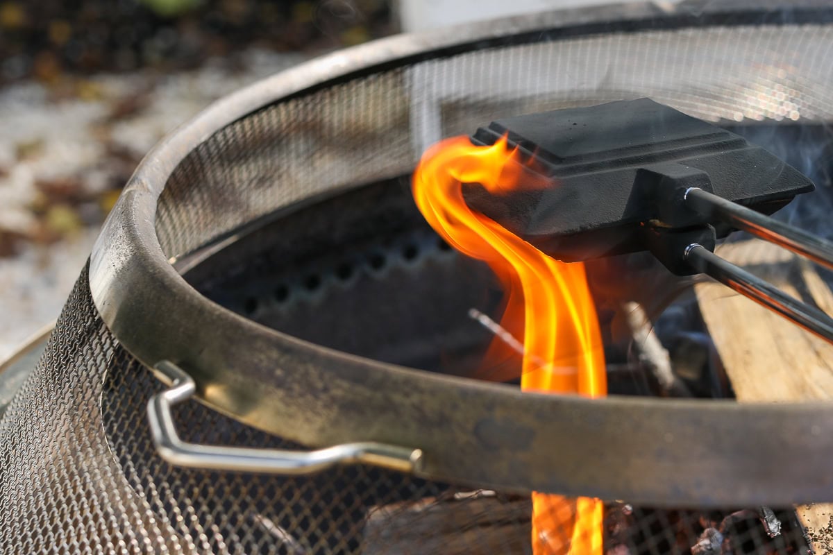 A pie iron with long handles is placed over an open flame in a metal mesh fire pit, reminiscent of making campfire grilled cheese on a cozy night under the stars.