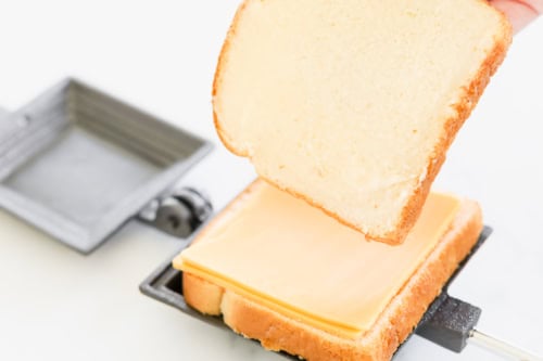 A slice of bread with butter is being placed on top of cheese and another slice in a sandwich press, creating the perfect campfire grilled cheese.