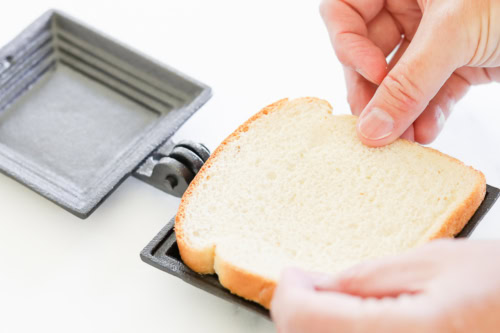 Hands place a slice of bread into a cast iron sandwich press on a white surface, ready to create the perfect campfire grilled cheese.